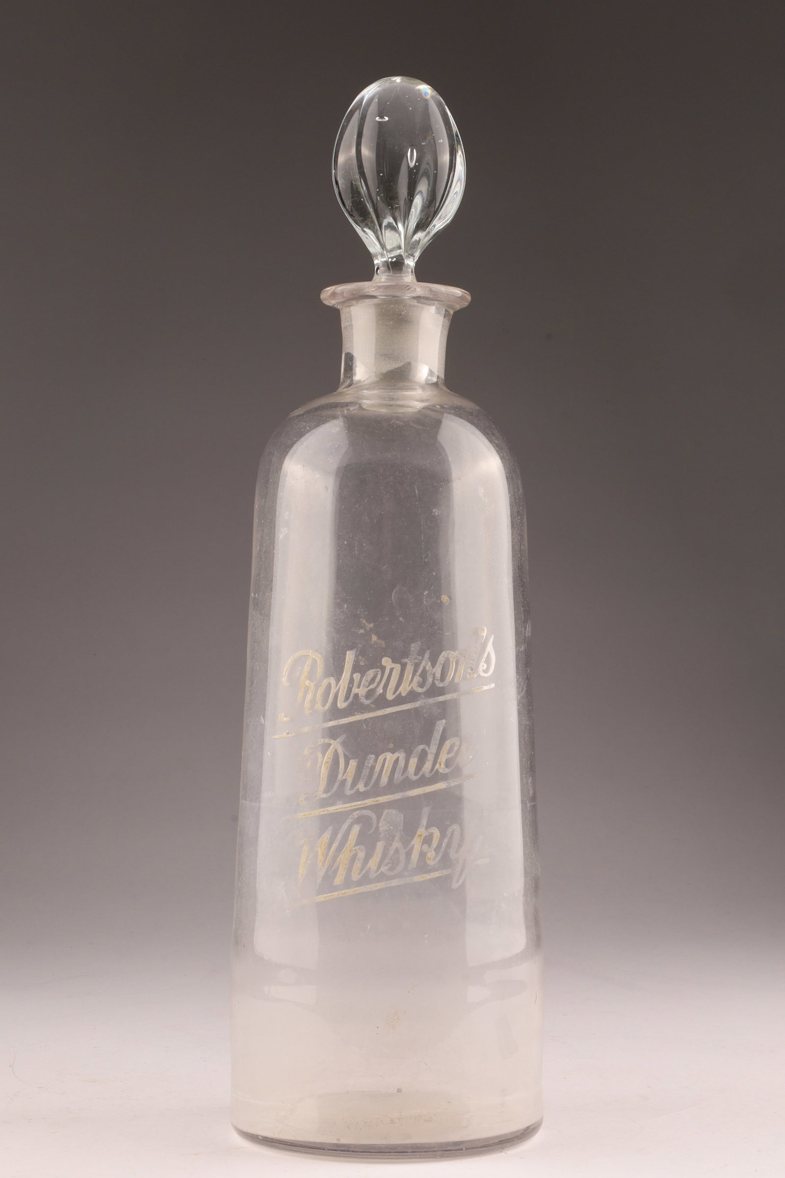Robertson’s Dundee Whisky Etched Glass Decanter with Stopper: A clear glass whisky decanter of cylindrical form, etched in script Robertson’s Dundee Whisky to the front. The bottle is fitted clear glass stopper, featuring a ground and frosted neck joint. The