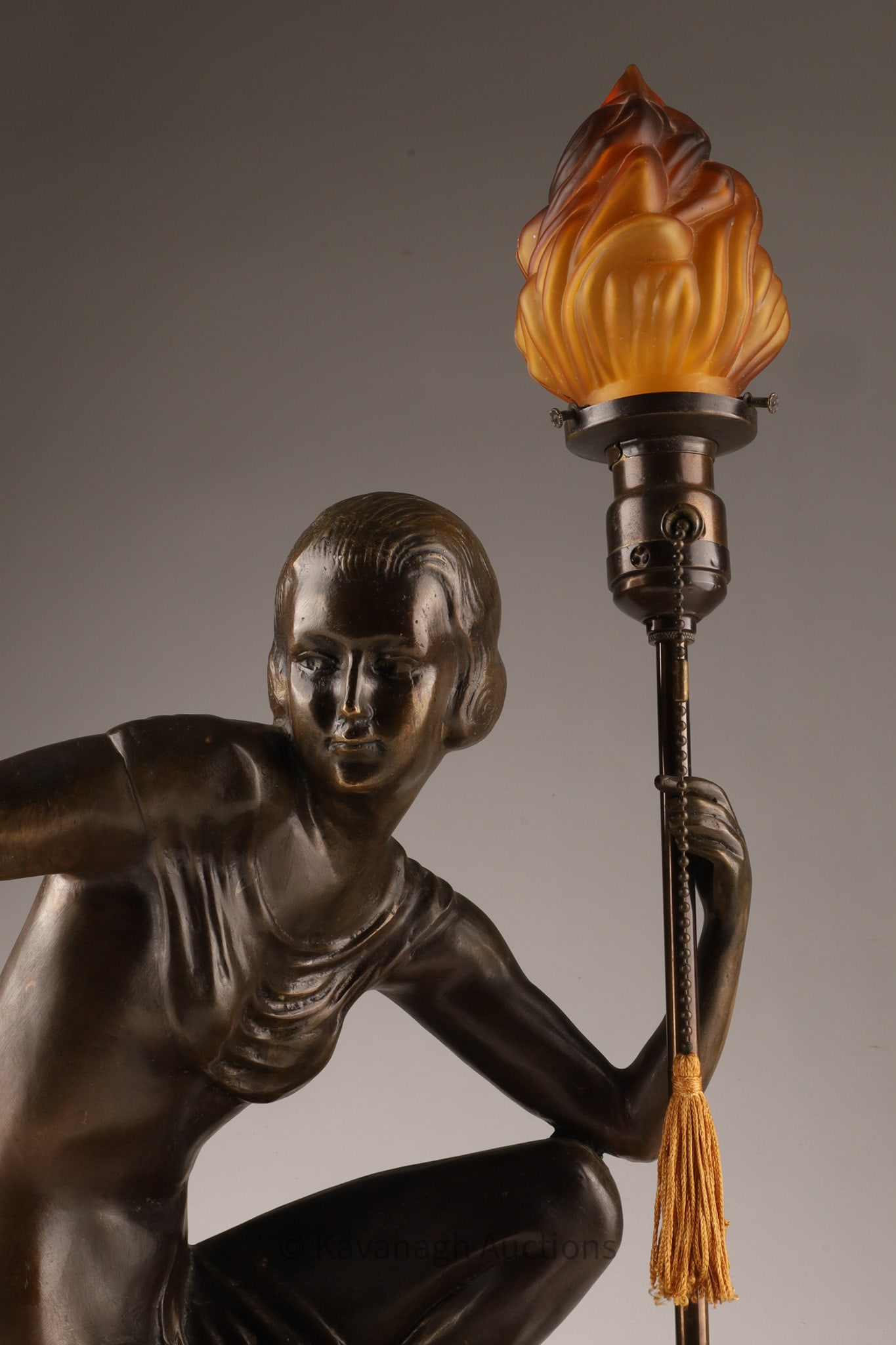 Art Deco Figural Table Lamp, Seated Female Nude with Torch: A sculptural Art Deco style figural table lamp depicting a seated female nude in a dynamic, asymmetrical pose, holding a torch-form light standard. Details Material: Bronze patina; stone base