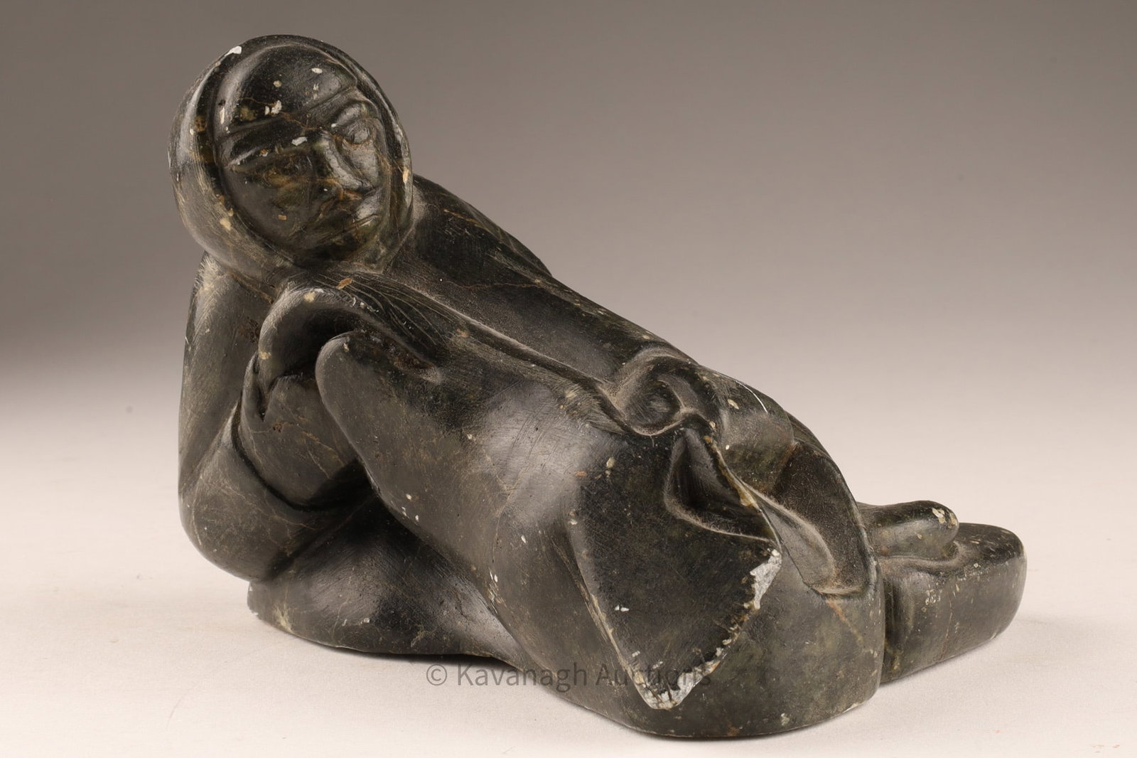 Signed Inuit Soapstone Sculpture of a Hunter with Seal, Arctic Canada: Inuit carved Soapstone sculpture depicting a reclining hunter figure holding a seal. The composition shows the figure lying on one side with head tilted slightly upward, the rounded face framed by a h