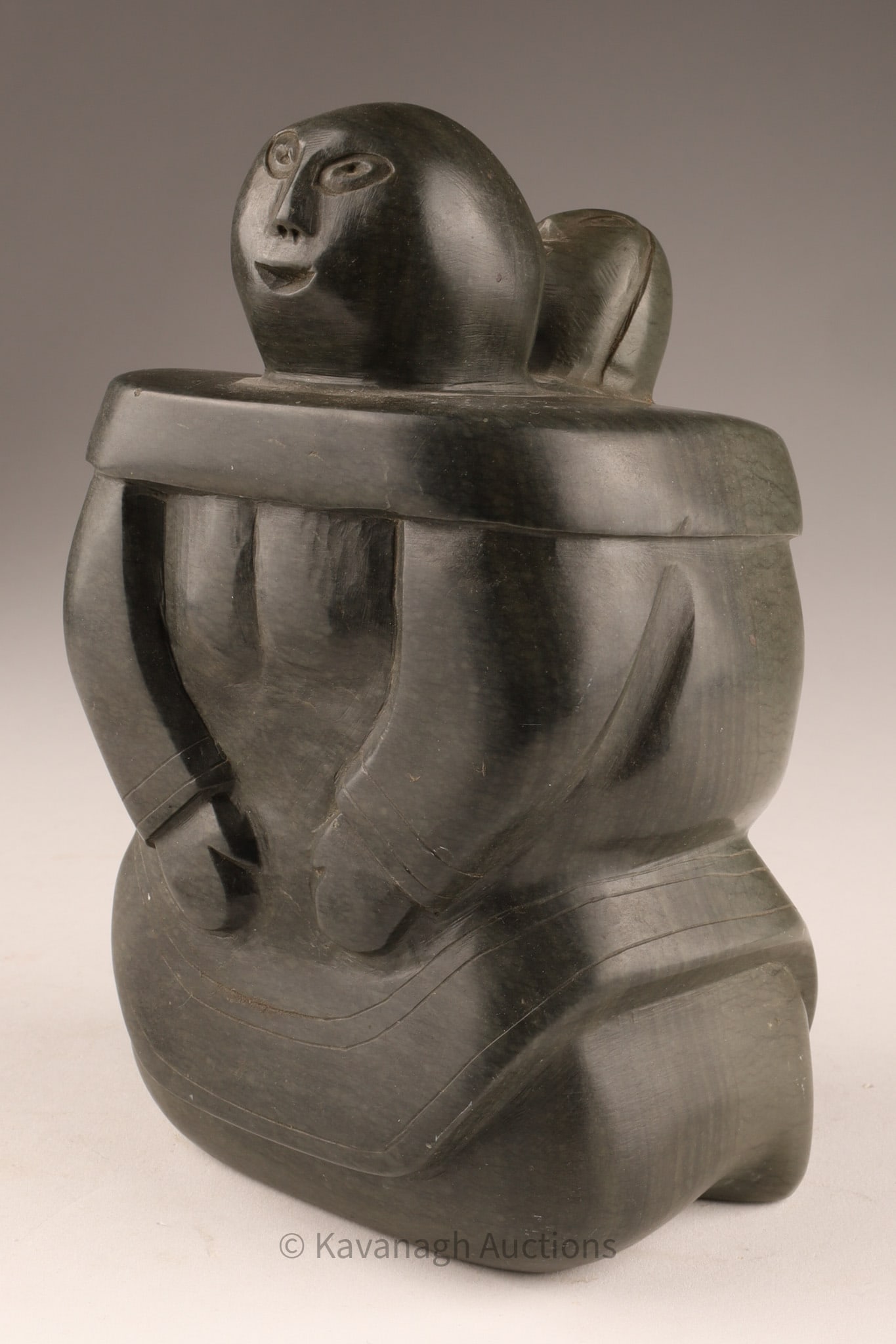 Signed Inuit Soapstone Mother and Child Sculpture: Inuit carved soapstone sculpture depicting a seated mother holding a child on her back. The underside bears a carved signature in Inuit syllabics along with an old inventory number and remnants