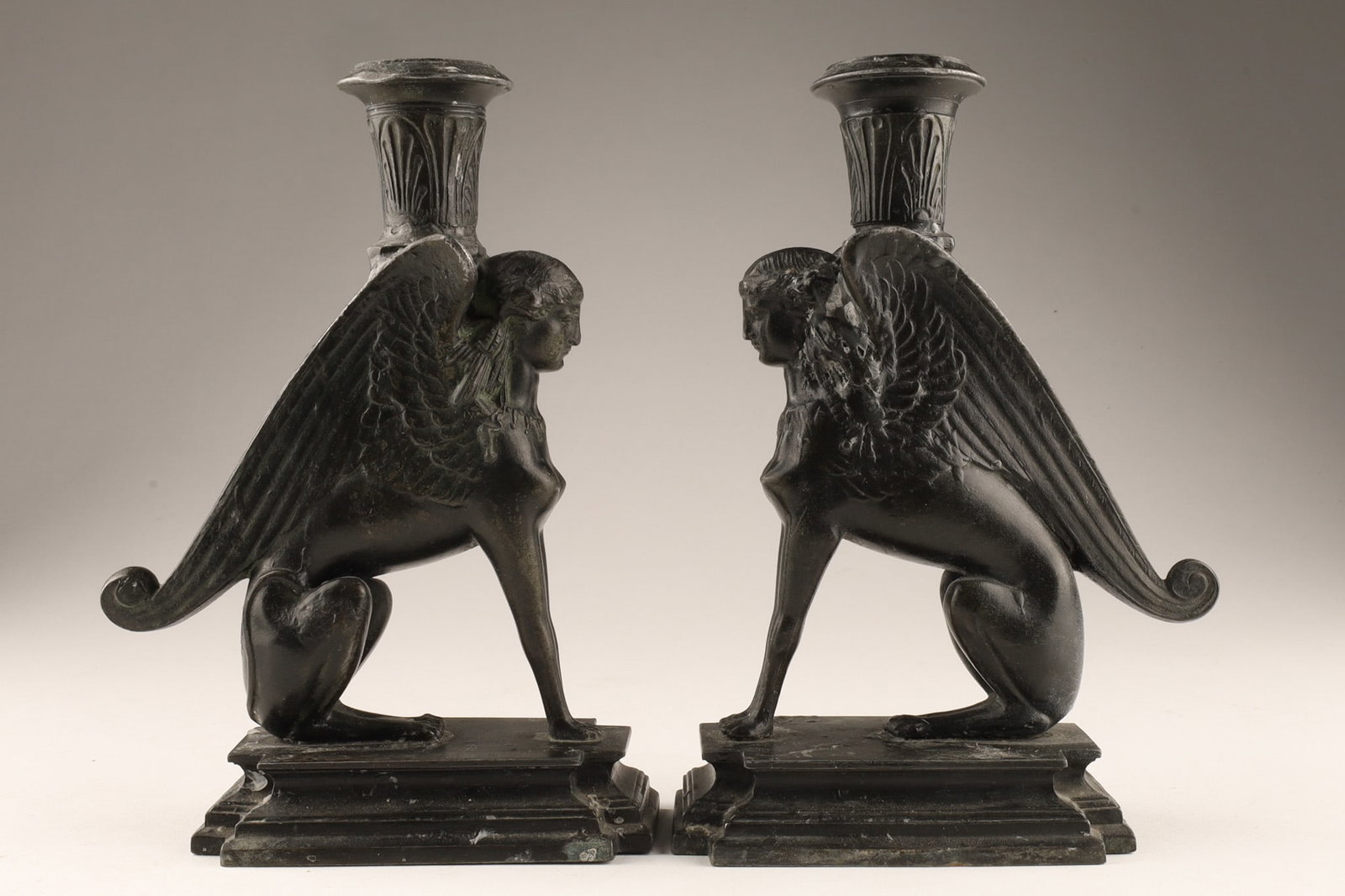 Pair of Art Deco Style Bronze Sphinx Mythological Candleholders: A pair of Egyptian Revival Style Sphinx candleholders, cast in bronze. Design: Each holder features a seated winged sphinx on a stepped rectangular base, with a candle nozzle (sconce) mounted on its h