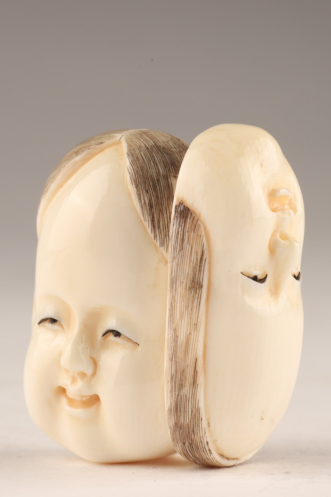 Japanese finely carved Netsuke of an Okame Otafuku Mask: Finely carved Japanese netsuke depicting the smiling face of Okame (Otafuku), shown with detailed coiffure and pierced himotoshi. Signed to the underside. Dimensions: 1.5 x 1.25 in. Note: THIS ITEM CA