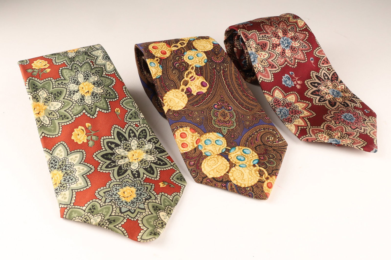 A Set of 3 Chanel Mens Silk Ties, Made in Italy: Set of 3 composed of one red base with large green and yellow floral medallions, one brown/Dark base with a jewelry print of gold chains and blue/yellow gems, and one deep burgundy with a floral star 