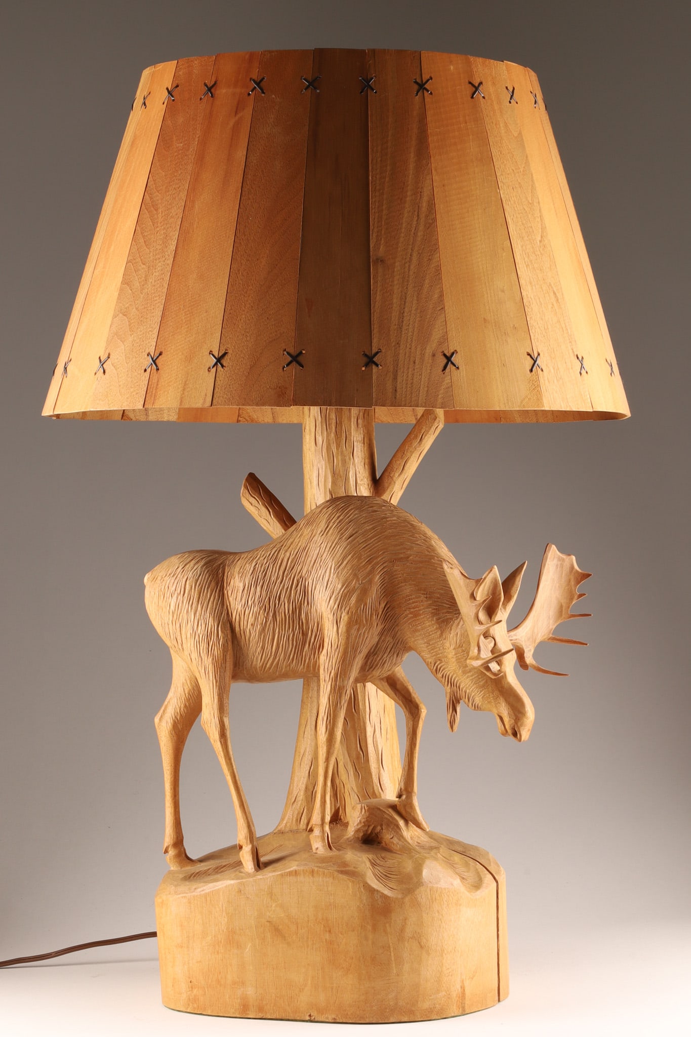 Andre Dube Folk Art Carved Table Lamp, Moose in Landscape: A folk art table lamp hand-carved by Canadian artist Andre Dube, carved out of a single piece of wood the moose stands with one leg resting on a raised area of ground, behind which a truncated tree fo