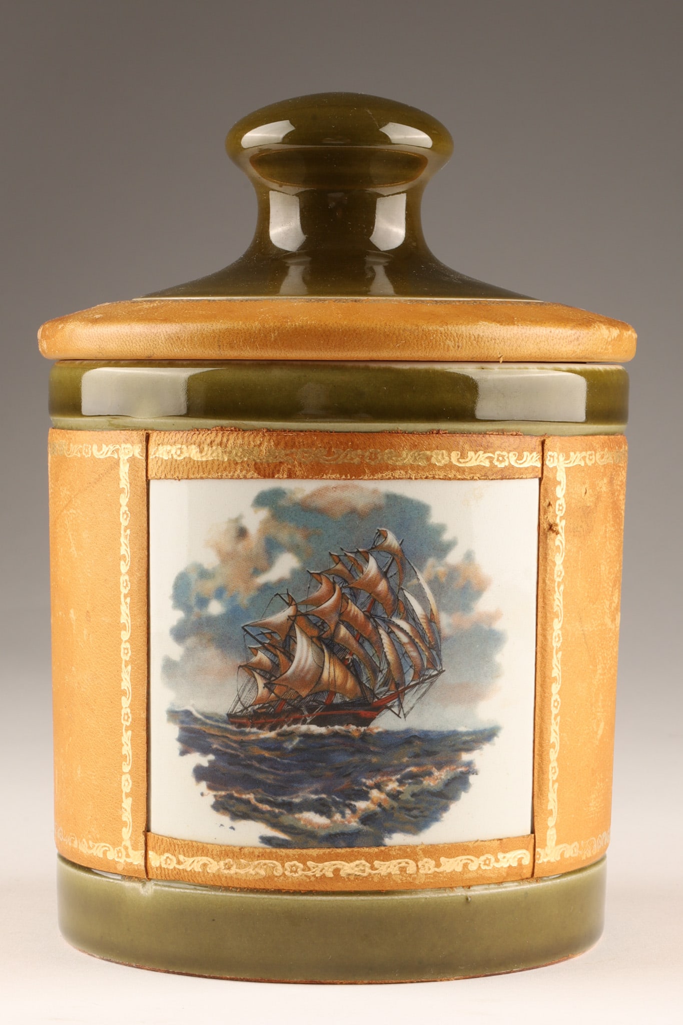 Italian Tobacco Humidor, Pottery Wrapped With Tooled Leather: An Italian ceramic tobacco humidor jar featuring a sailing ship motif with an olive green glaze and lighter tan leather wrapping. Marked on underside, Genuine Leather Italy. Dimensions: 6.5 inches H x