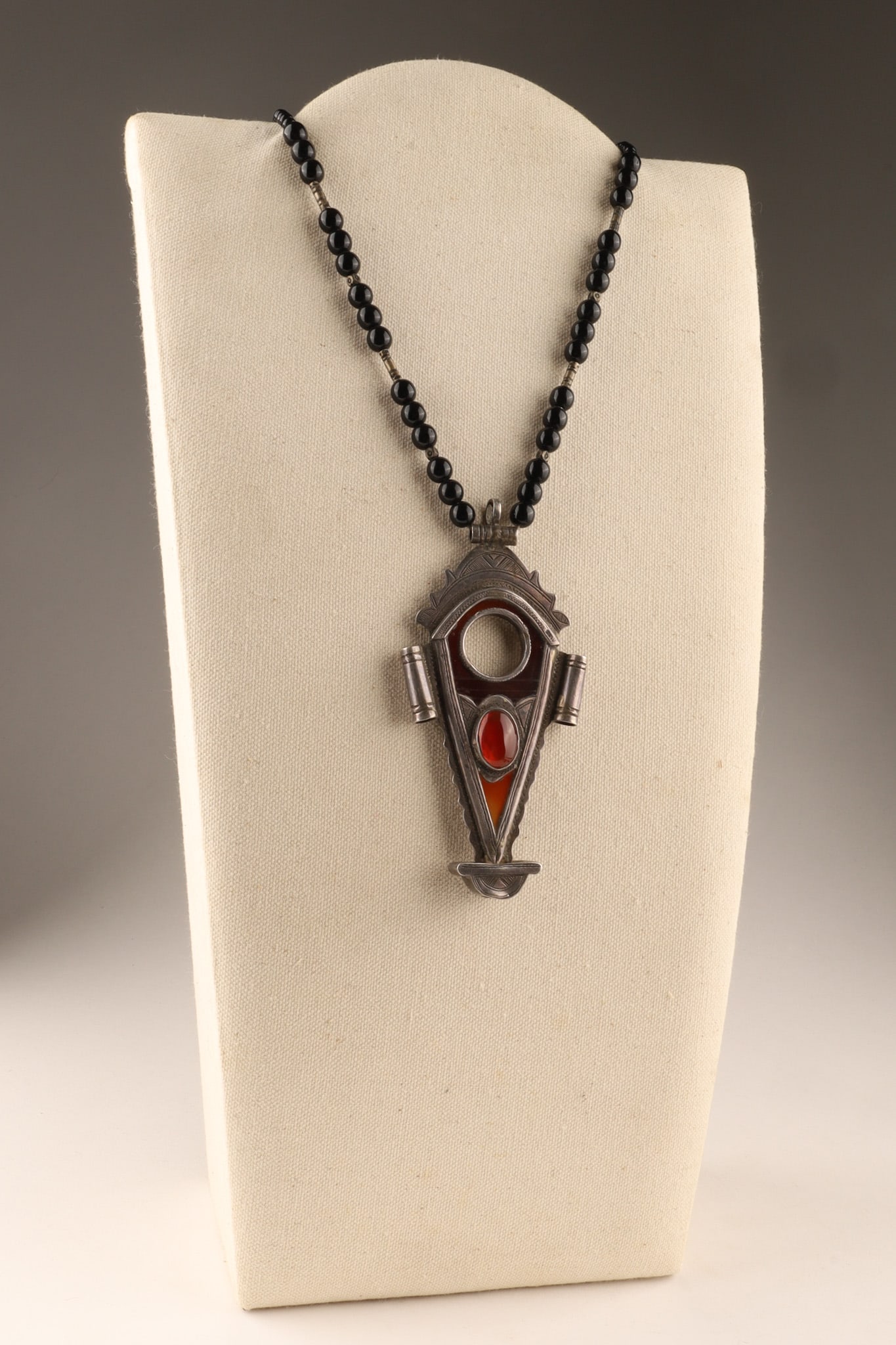 North African Silver Tuareg Amulet Necklace with Carnelian Inlay and Onyx Beads: North African Silver Tuareg Amulet Necklace with Carnelian Inlay and Onyx Beads, Known as a Tanfouk or Talhakimt. The central circular opening acts as an architectural focal point, housing a rich red 