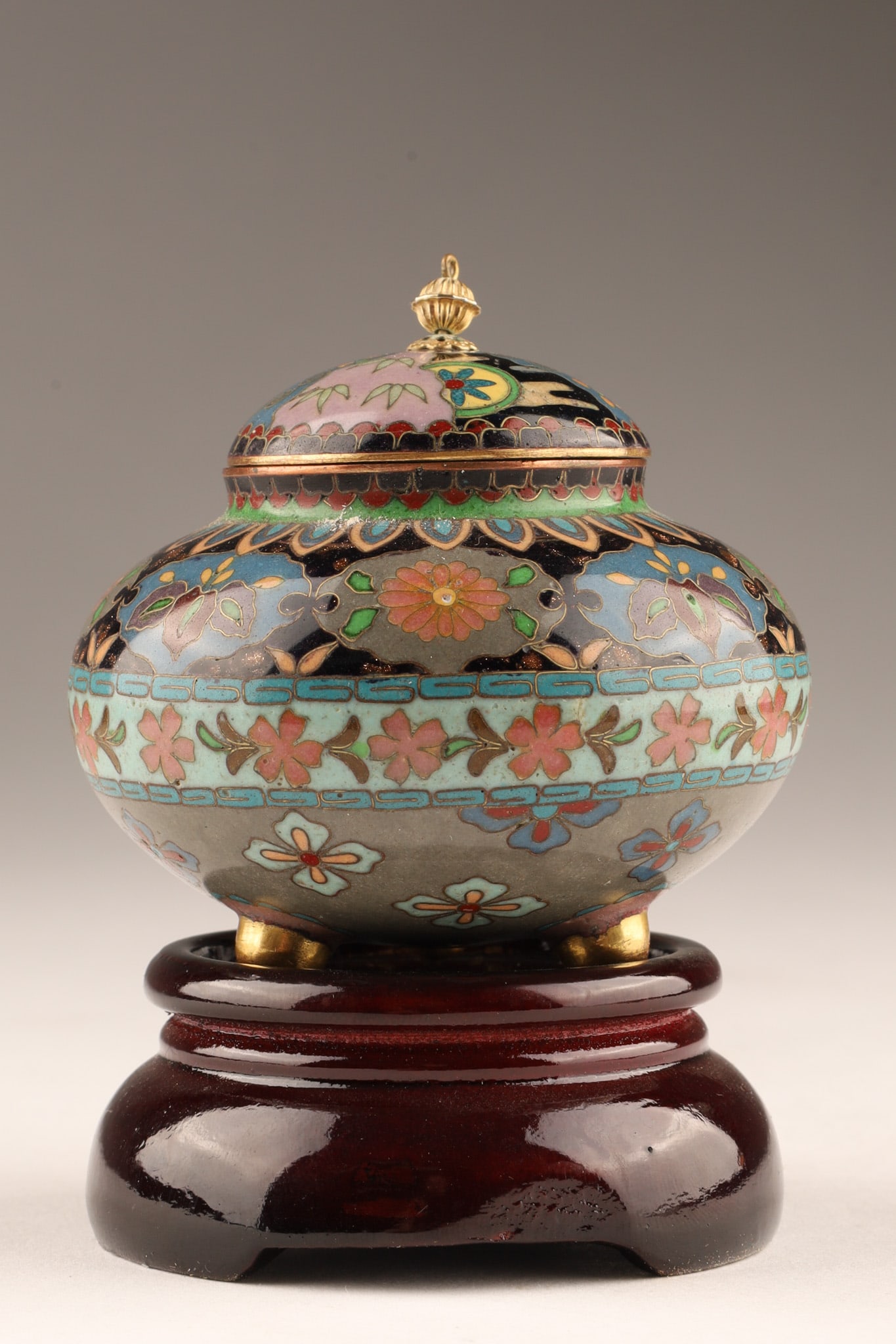 Antique Japanese Small Cloisonne Lidded Tripod Jar on Stand: An antique Japanese small cloisonne lidded jar featuring floral motifs and tripod legs, upon a modern wood stand. Jar measures 2.5 inches H x 2.75 W, on stand measures 3.5 inches H.