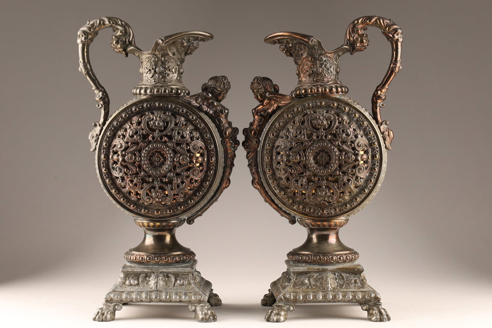 Pair of Antique Cast Metal Figural Mantel Ewers Cherubs and Faces: Pair of Victorian or Renaissance Revival antique cast metal decorative figurative mantel ewers or urns, the round body set with a cherub and faces, set on square raised bases with lion paw feet, Europ