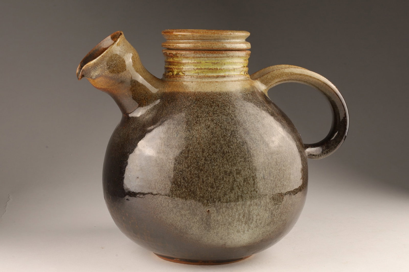 Mayta Markson (Canadian, 20th Century) Studio Pottery Teapot / Pouring Vessel: A mid-century stoneware teapot featuring a spherical wheel-thrown body, an angled tubular pouring spout, and an over-slung strap handle with a rich, dark metallic reduction glaze that transitions into