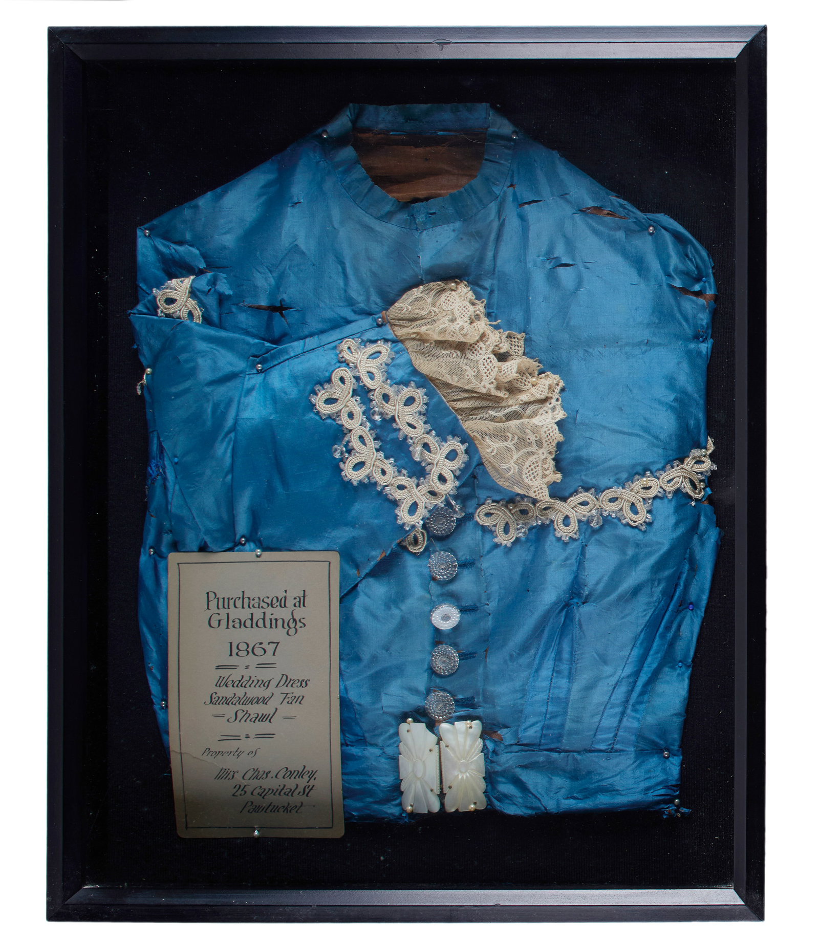 1867 Gladding's Department Store Wedding Dress: Antique blue wedding dress with applique and lace detailing and what appears to be a mother-of-pearl clasp or buckle, framed. 1867. Included in the frame with a note reading: "Purchased at Gladdings,