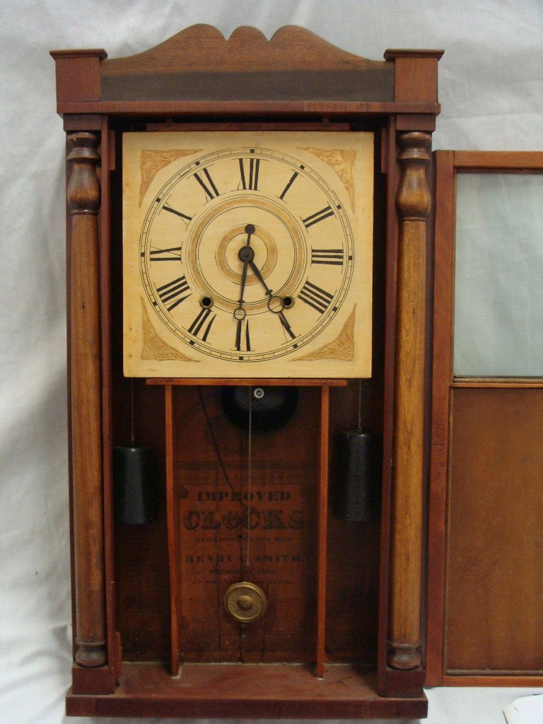 Improved Henry C. Smith Wall Shelf Pendulum Clock: 27 1/2" tall, 31" with topper, 16 1/2" wide, 4 1/2" deep. Appears to have a replacement minute hand which is loose, door is in good shape, hinges are in need of repair, replacement mirror is cracked.