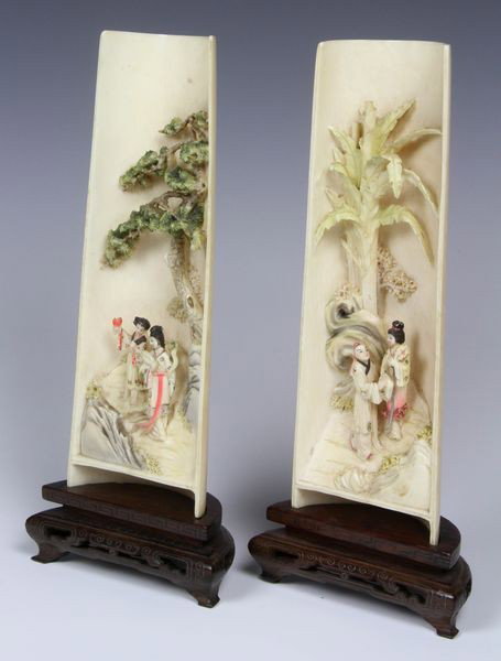 Two (2) Chinese 19th/ 20th C. Carved Ivory Sleeve: A pair of ivory sleeve dioramas, China, 19th/ 20th century, each of half cylinder shape carved in relief with figures at leisure beneath trees, decorated with paint, on carved wood stand, 7"h.
