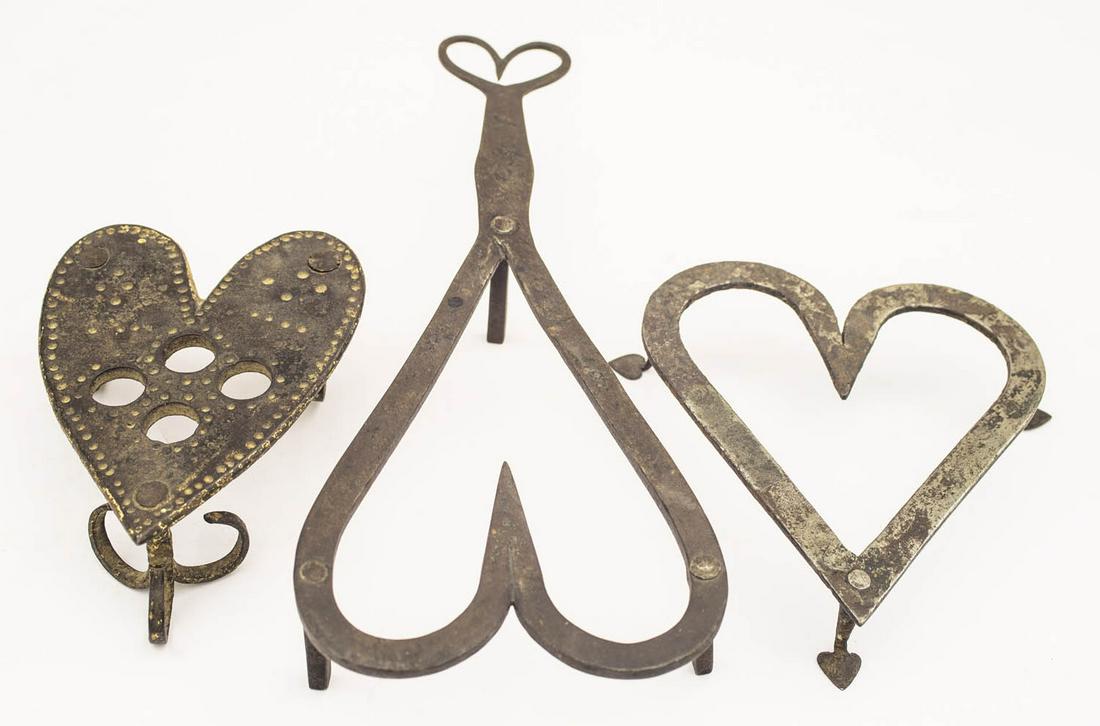 THREE EARLY WROUGHT IRON TRIVETS Each with heart motif,: THREE EARLY WROUGHT IRON TRIVETS Each with heart motif, one with long (11") handle ending in a heart, one with finely shaped spade feet, 5 1/2" l, and one with punch decorated solid top (with four hol