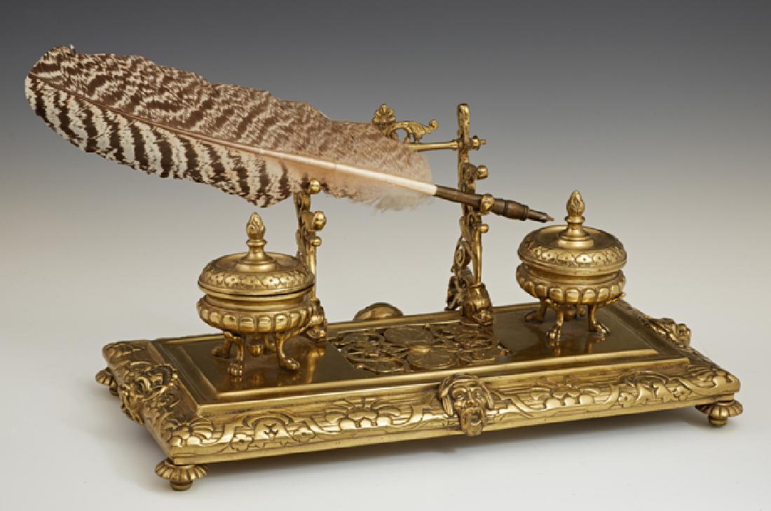 French Gilt Brass Inkwell Pen Tray, late 19th c., with: French Gilt Brass Inkwell Pen Tray, late 19th c., with a rear pen rack behind two lidded urn form inkwells, on a pierced relief base with relief faces centering the four sides, on disc feet together w