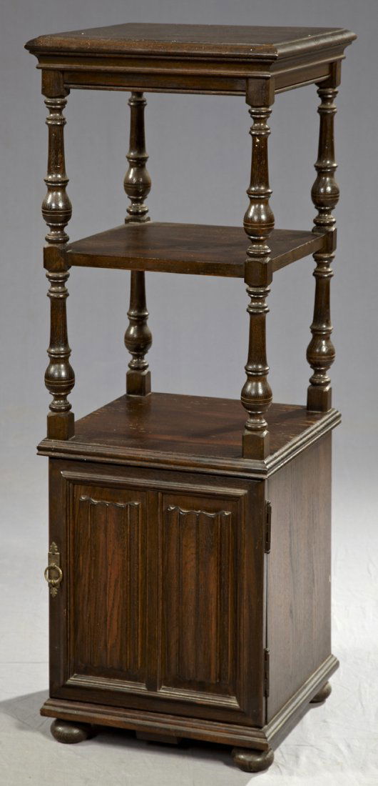 English Carved Oak Tall Pedestal, c. 1920, the ro: English Carved Oak Tall Pedestal, c. 1920, the rounded edge top on turned supports to a middle shelf, on turned supports, to a lower shelf over a cupboard door with relief linen fold decoration, on tu