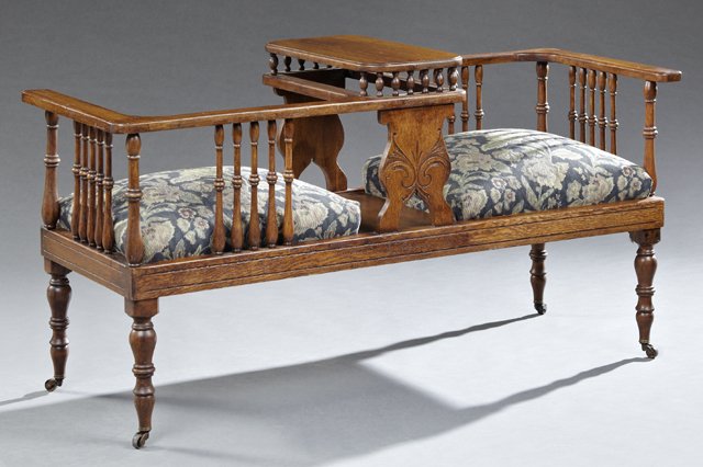 Edwardian Carved Oak Tete-a-Tete, c. 1910, the two: Edwardian Carved Oak Tete-a-Tete, c. 1910, the two opposing slip seats with spindled backs and sides centered by an open shelf, on ring turned tapered legs to casters, H.- 27 1/2 in., W.- 52 1/2 in., 