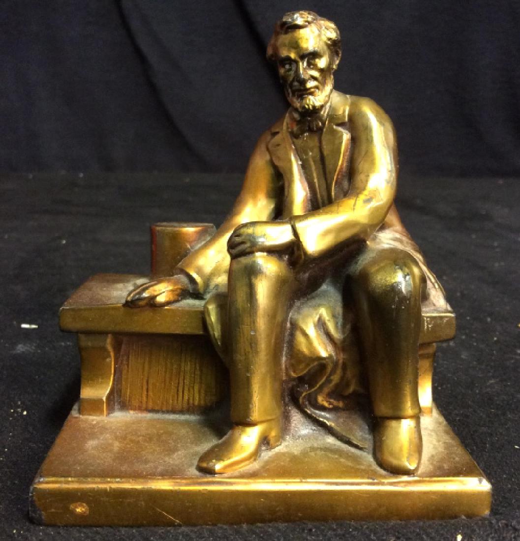 Brass Abraham Lincoln Figurine: Abraham Lincoln figurine is gold toned brass metal. Statue of Lincoln is sitting on a bench next to his hat. Base is rectangular shaped. Contains slight discoloring, bottom of base contains rust. Meas