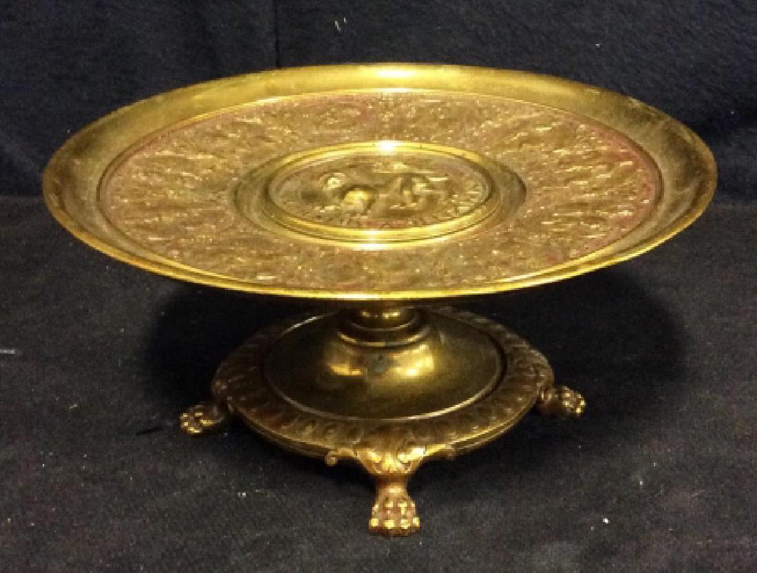Brass Toned Table Top Accessory: Claw footed pedestal dish with raised filigree applied design, top with center lion detail, and an embossed rim design of military figures on horseback, some surface wear including discoloring and scr