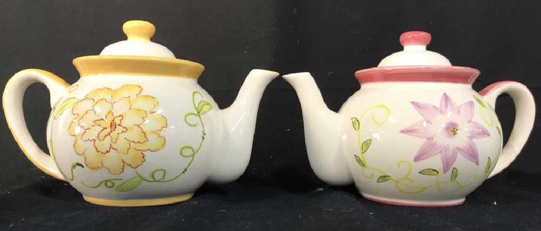 Lot 2 JOHN HENRY Porcelain Teapots: Labels on underside read JOHN HENRY. One has pink toned painted accents, the other has yellow toned painted accents. Each has painted floral, leaf, and vine detail. Measures approx 9 inches long, 6 in