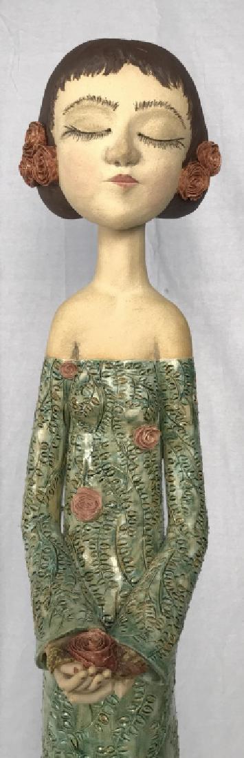 MEL REA Pink Roses Sculpture: A Ceramic Sculpture by Mel REA titled Pink Roses, sculpture depicts a ceramic figure of a standing woman, with eyes closed, roses in her hair, holding a rose bud in her hands and wearing a green vine 
