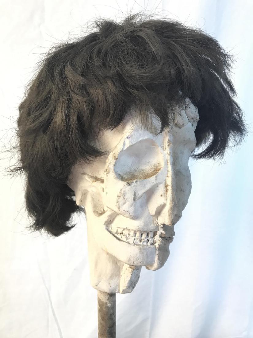 Plaster Skull Figure W Wooden Base: Piece appears to be plaster. Base appears to be wooden. Contains brunette wig on top. Skull sculpture, plaster sculpture, Sculptural art. Measures approx 23 inches tall 11.5 inches wide, 15 inches dee