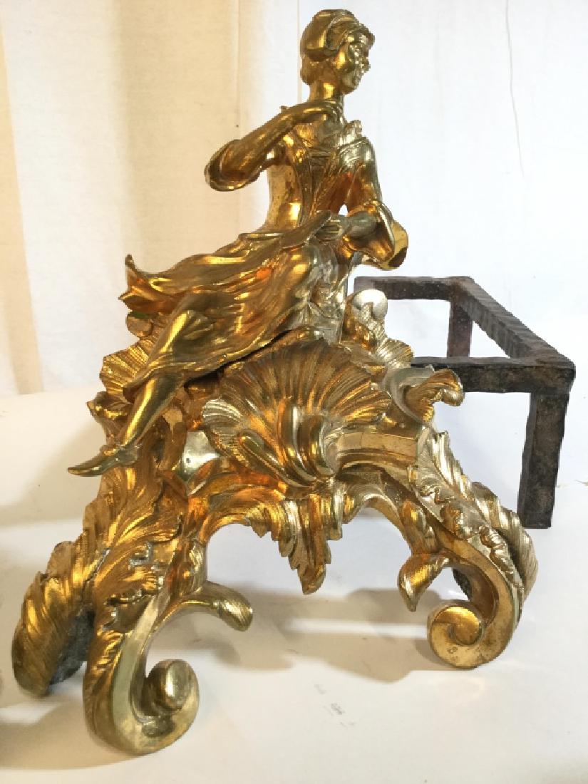 Pair Gold Toned Brass & Iron Andirons: Andirons are gold toned. Appear to be solid brass. One depicts a male figure, the other depicts a female figure. Each measures approx 12 inches tall, 17 inches long, 11 inches deep. Andirons, Brass an