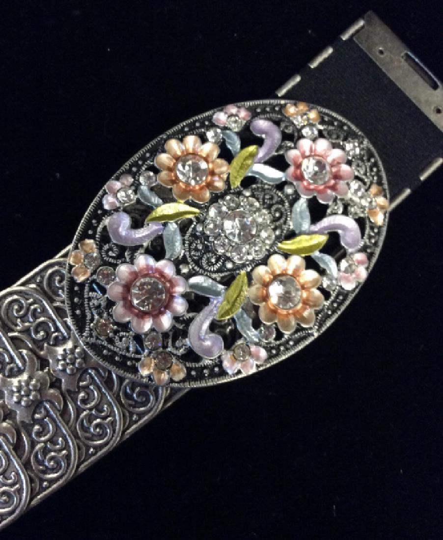 Belt W Bejeweled Floral buckle: Stretch fabric base,, Silver toned metal link belt with raised curling line filigree motif, rhinestone details buckle with floral forms and multi toned enamel details. Measures approx 30 in from end t