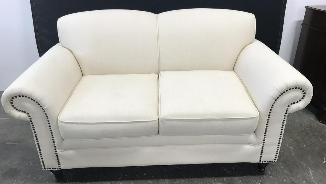 Upholstered Couch W Tack Detailing: Off-White tone Fabric Upholstered Couch With tack detailing. Some surface wear of couch, including blemishes. Back leg needs reaffixing. Measures approx 34 inches in height by 64 inches in width by 33