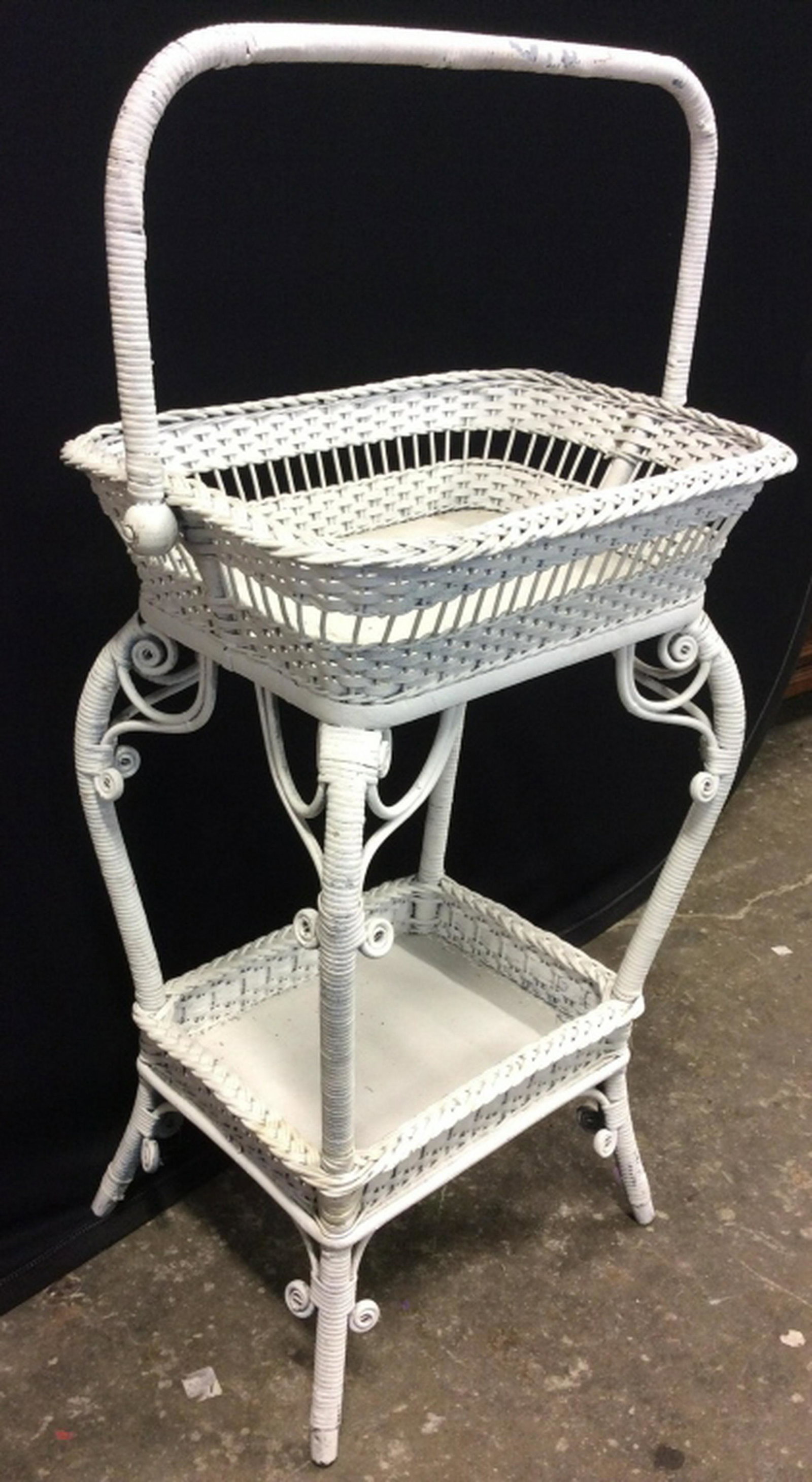 White Toned Wicker Basket Table: Piece is white toned & wicker. Table top has basket like structure with handle. Piece also has lower shelf like area. Piece measures approx 28 inches tall 20 inches wide, 12 inches deep. whicker. home