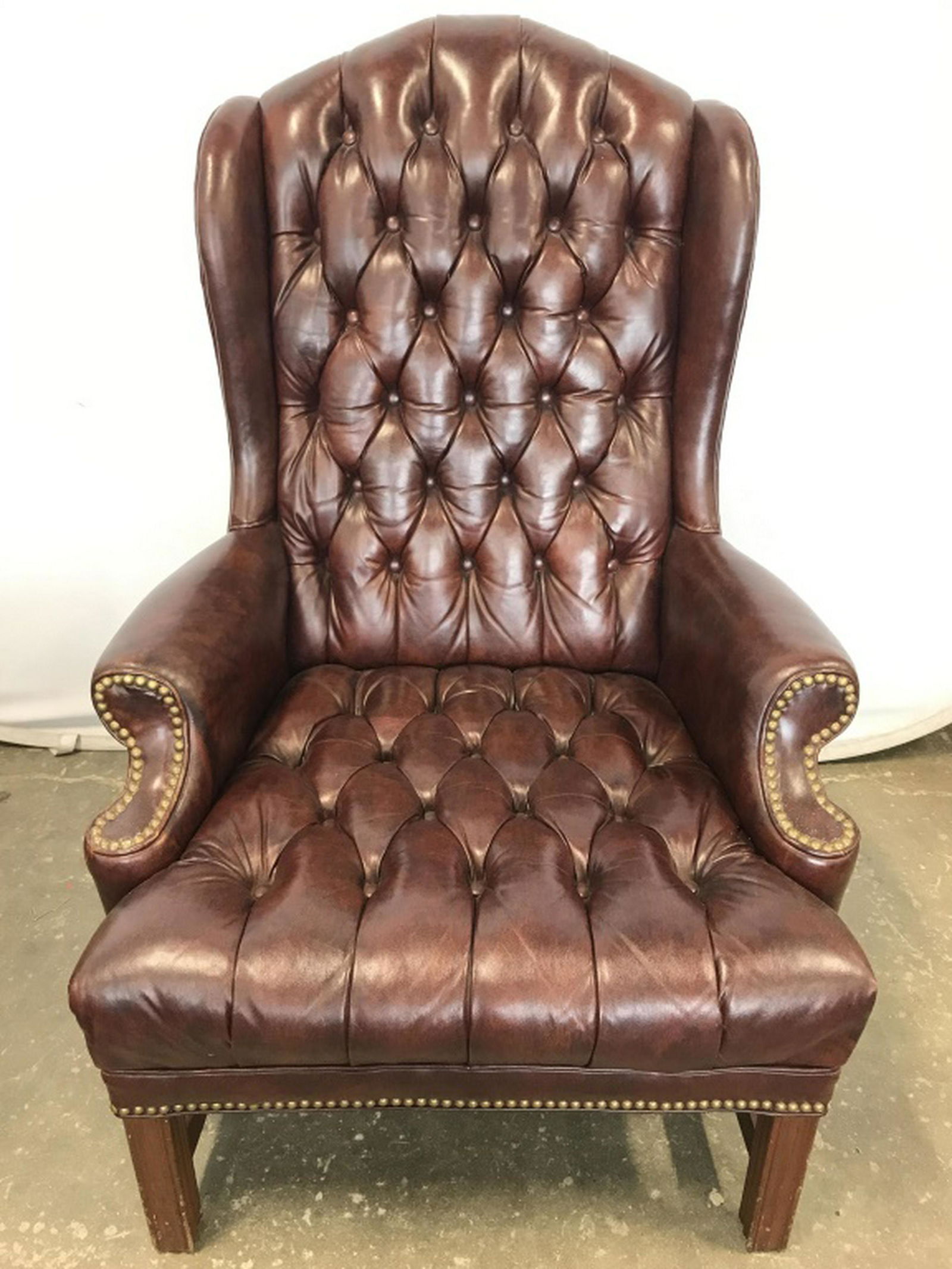 Tufted Leather Wingback Arm Chair: Chair has a burgundy brown tone. Piece is Tufted with winged backrest. Contains gold toned studded detail. Piece also has carved wooden legs. Piece measures approx 44 inches tall 29 inches wide 35 inc