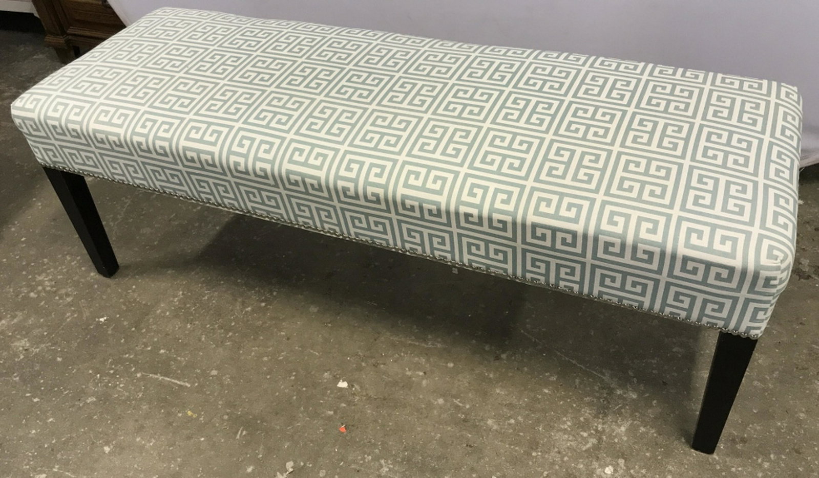 Bench W Geometric Pattern Upholstery Long bench with: Long bench with geometric sea foam green and off-white tone fabric upholstery with silver tack trim details. Bench sits atop black tone wood legs. Measures approx 56 inches in width by 19 inches in he