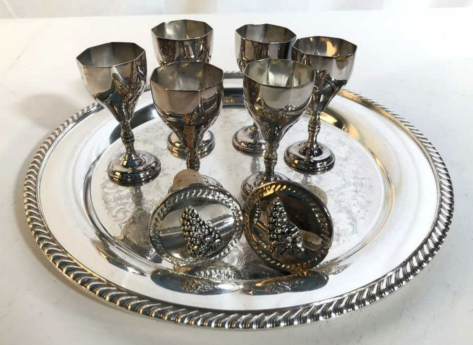 Silver Pl Tray w 6 Cordial Cups Wine Stoppers Tray is: Tray is etched with rope twist border marked underside for WM Rogers, diameter app 12 inches, Cordial goblets stem ware each measure ht 3.5 inches, two silver wine Stoppers with raised grape motif