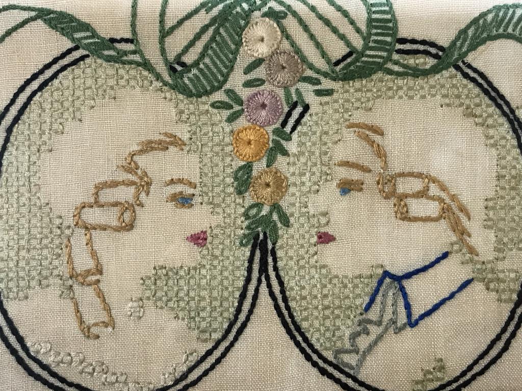 Vintage Couple’s Sampler: Sampler features man and woman couple Motif with wreath border. Sampler is set behind glass in wood frame. Frame measures approx 6.5 inches in height by 13.4 inches in width. Sampler has surface wear 