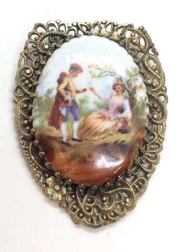 Porcelain plaque Pendant: Brass toned metal pendant with openwork filigree designs, oval porcelain plaque with romantic scene of Victorian esque couple in wooded park scene. Measures approx 2 1/2 in by 1 1/2 in, women’s vint