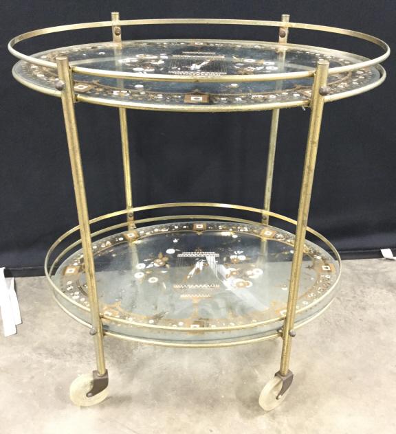 Vintage Metal & Glass Bi Level Tea Cart: Tea cart is metal and glass. Contains white and gold toned floral, leaf, bird, and geometric detail. Piece has 4 wheels. Each level has an oval shape. Piece measures approx 27 inches tall 25 inches lo