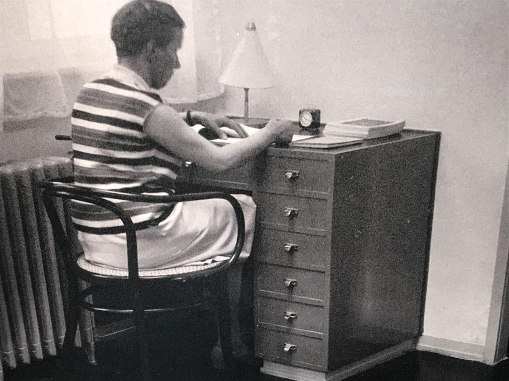 Framed B&W Lithograph Of Vintage Photograph: Vintage black & white tone lithograph depicting photograph of figure working desk while sitting on a bentwood chair. Possibly an offset lithograph. Print is set behind plexiglass in white tone wood fr