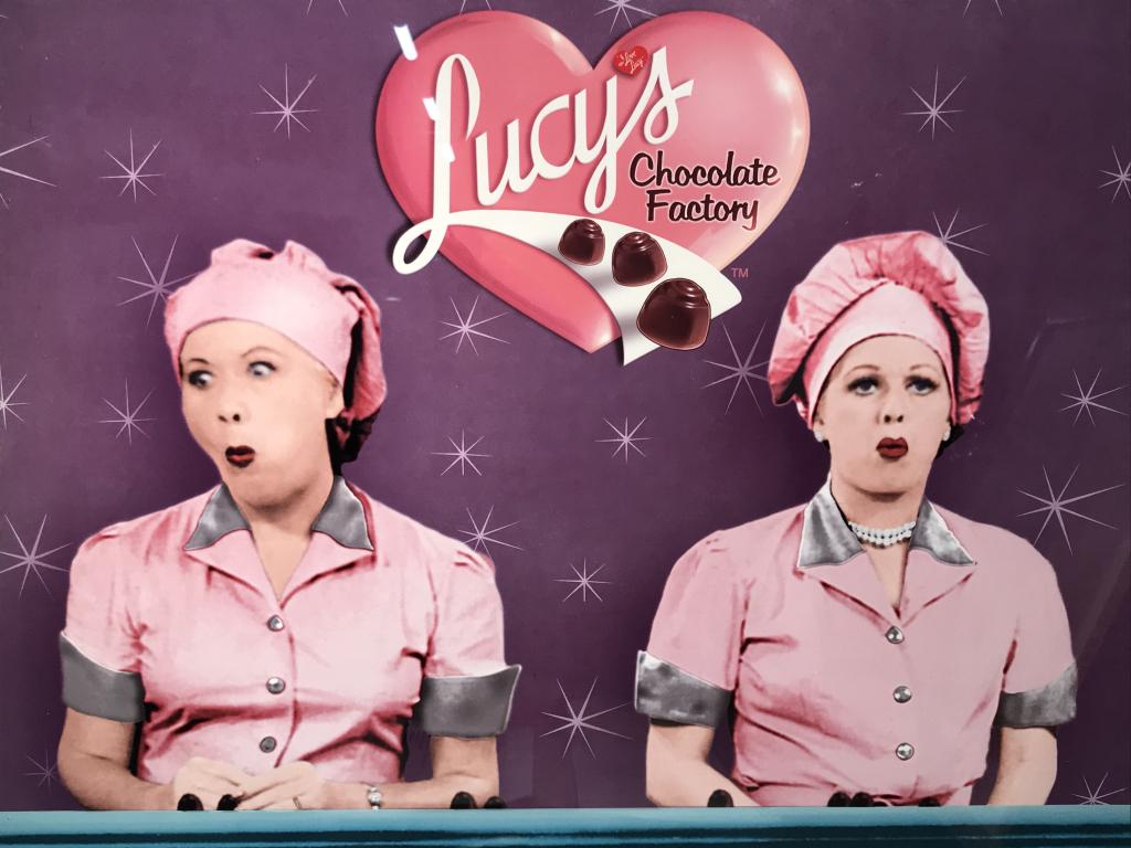 Framed I Love Lucy Chocolate Factory Poster: Framed color poster print of I Love Lucy’s iconic Chocolate Factory Scene. Poster is set behind glass in white tone coated wood frame. Measures approximately 21 inches in height by 27.25 inches in w