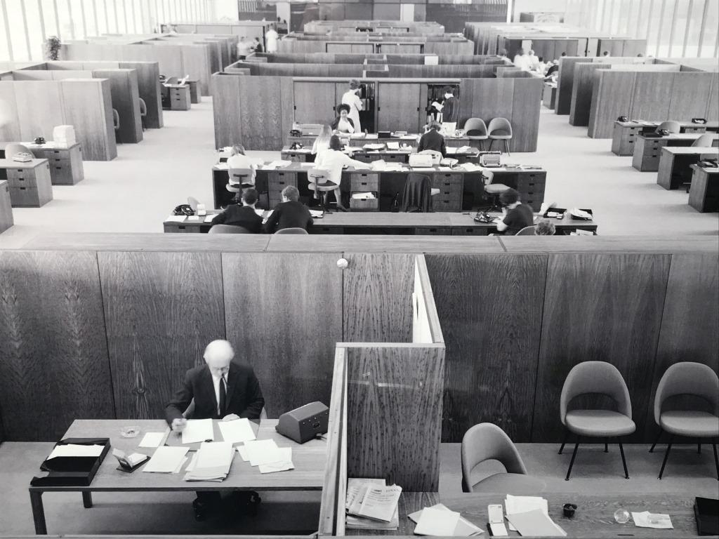 Framed B&W Lithograph, Vintage Office Interior: Vintage black & white tone lithograph depicting architectural interior of office building interior with figures working. Possibly an offset lithograph. Print is set behind plexiglass in white tone woo