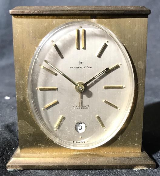 HAMILTON Gold Toned Desk Clock: Desk. clock appears to be brass. Piece is gold toned. Piece has oval shaped clock face. Clock face reads HAMILTON. Desk clock, tabletop accessories, desktop accessories. Measures approx 3 inches tall 