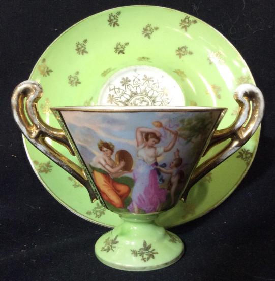 VICTORIA Austria Teacup and Saucer: Lime green toned on white toned porcelain / China. Marked on underside VICTORIA AUSTRIA. Dual Handled design, with gold toned floral design and trim, and romantic garden scene with robed women and Cup