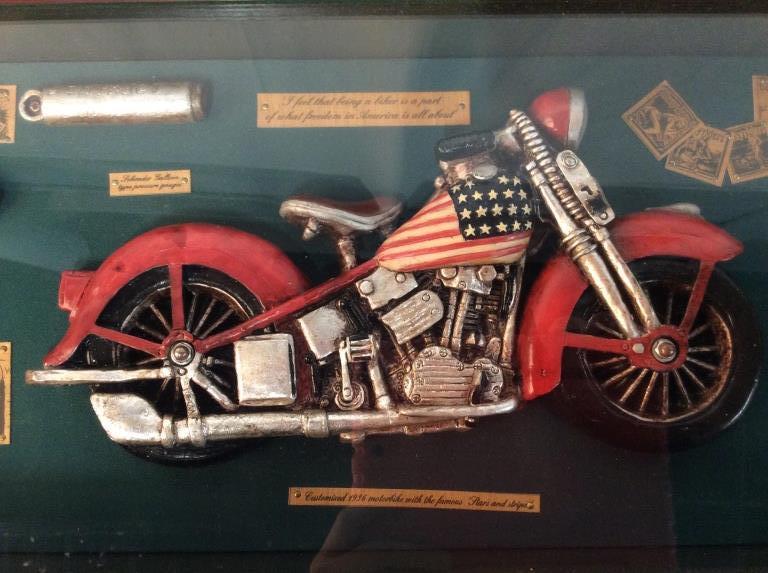 Motorcycle Americana Shadowbox Display: shadow Box with dimensional plaques of customized 1936 motorcycle with american flag motif, leather jacket with quote “ love to ride “ and riding goggles. Plaques unknown material possibly wood or