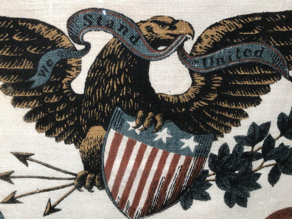 Framed Americana Stamp Dyed Textile: Framed Americana stamp dyed textile depicting eagle with shield carrying banner that reads ‘We a Stand United.’ Textile is folded stylistically, set behind glass in wood frame. Measures approx 14.