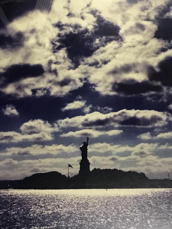 Framed Photographic Print, Sun Shining on Liberty: Framed photographic print of the sun shining down on the Statue of Liberty in NYC. Artwork is matte and set behind plexiglass in white tone wood frame. Measures approx 39 inches in height by 29 inches