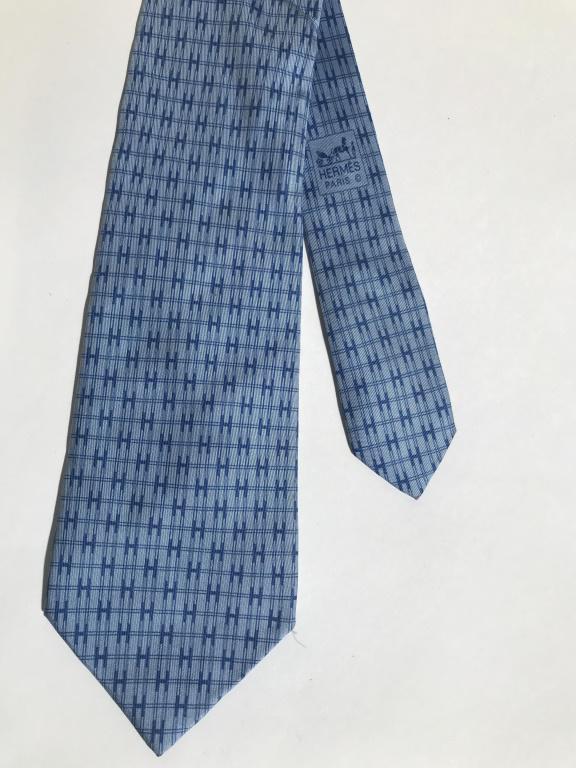 HERMÉS PARIS Silk Men’s Necktie: Hermés Paris necktie, the tie is 100% silk. Pattern covering the tie depicts a series of H’s and other geometric shapes. The tie is colored in blue tones, with a matching red lining. The piece meas