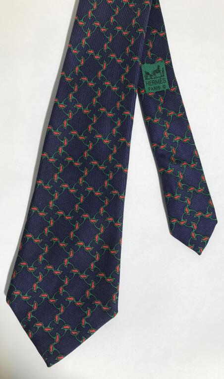 HERMÉS PARIS Silk Men’s Necktie: Hermés Paris necktie, the tie is 100% silk. Pattern covering the tie depicts a series stirrup shaped objects linked together. The tie is colored in blue tones, green and red, with a matching blue lin