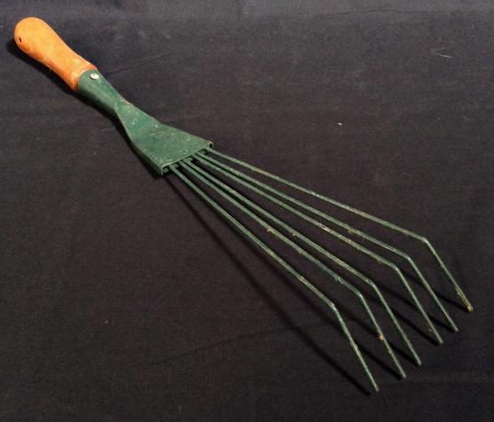 Handheld Gardening Rake: This piece is a handheld gardening rake. The piece is colored in green tones and is fastened to a wooden handle. The piece measures approx 19.5 inches Long, and 3.5 inches W. Some wear is present due 
