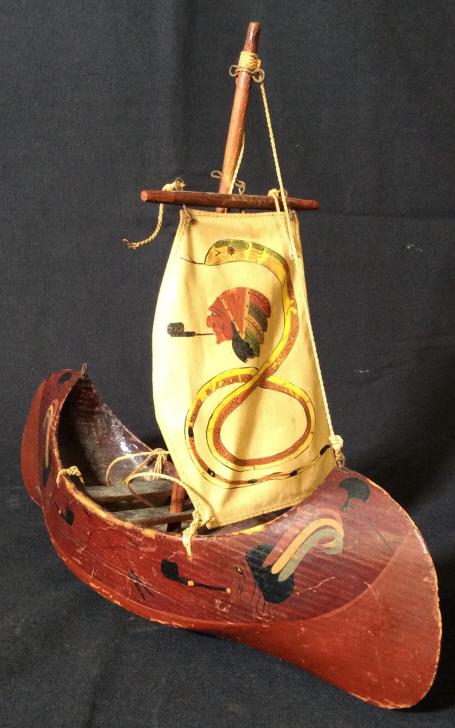 Vintage Wooden Native American Sailboat: This piece is a wooden sailboat, the piece is vintage and colored in crimson tones, and is hand painted. The sail is decorated with the face of a male figure and a snake. The piece measures approx 9.3