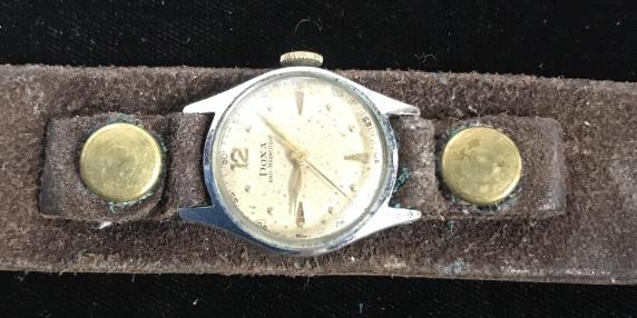 Vintage DOXA Anti-Magnetic 8 in. Wristwatch: Brown suede band, 1 in wide. Watch case sits on top of band. Has brass snap button accents, brass dial and brass buckle. Backside on watch impressed with numbers ‘5127733’. Scratched glass and wor