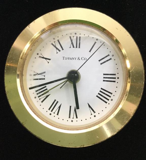 TIFFANY & CO Gold Toned Brass Desk Clock: Clock is gold toned possibly brass with white tone clock face. Clock face reads TIFFANY & CO. PIECE measures approx 2.75 inches tall 1.15 inches deep. reverse side reads MADE IN GERMANY. Contains some