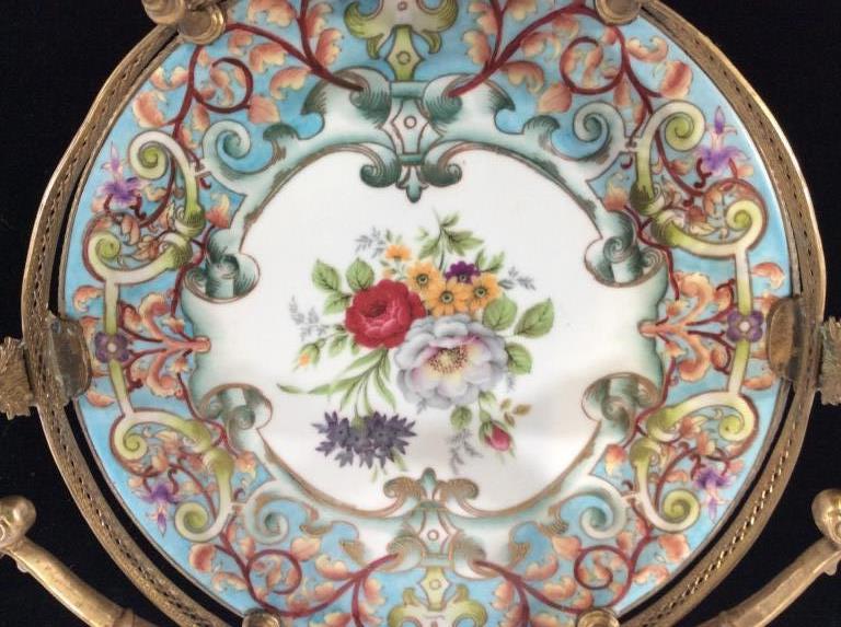 Victorian Brass And Porcelain Footed Serving Tray: Hand painted porcelain over glaze, center floral design, scrolls and gilt outlining. Embossed brass pillars flank the Pierced brass rim. Carry Handle swivels side to side. Vintage piece. Fairly heavy.